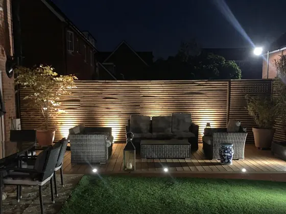 A lit deck at night with garden furniture, uplighters washing the slatted fence behind